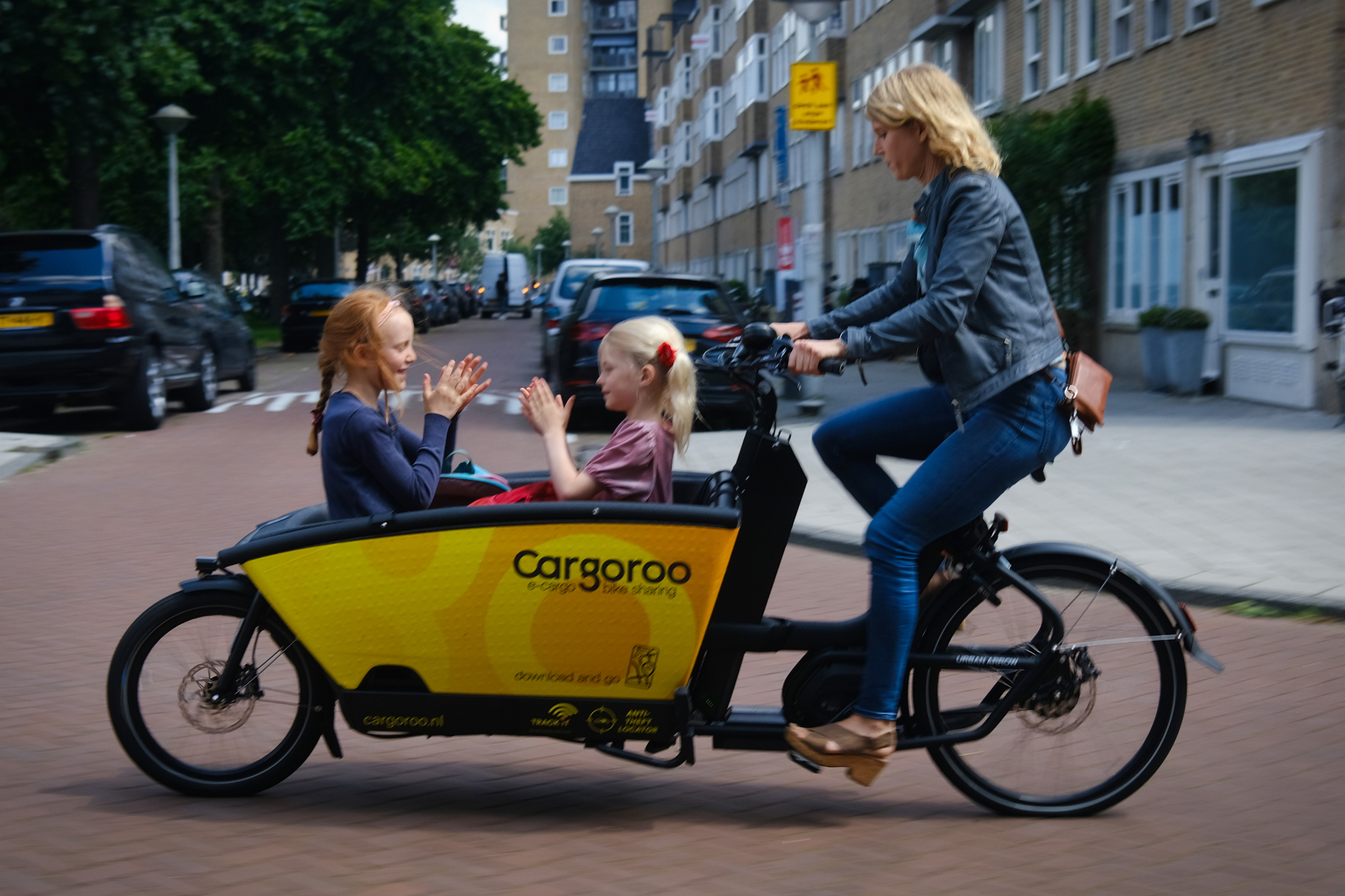 A parent rides two children in a yellow Cargoroo e-cargo bike through an Amsterdam street